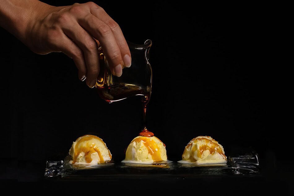 За какво се използва олио от трюфели? 3 Ръка налива сироп върху три топки сладолед върху стъклен съд, подчертавайки изисканото използване на тъмен фон.